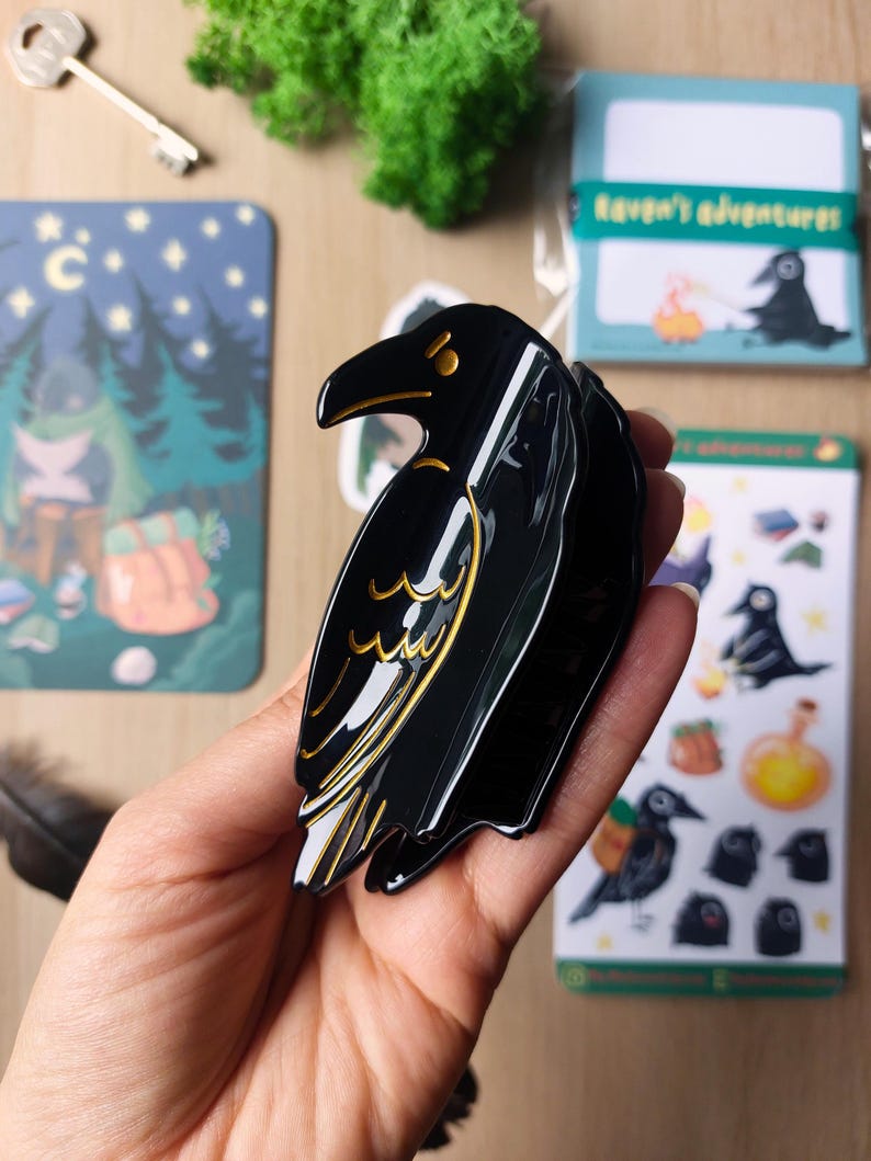 K&ouml;nnte beinhalten: Eine schwarze Haarspange in Form eines Raben mit goldfarbenen Linien, die den Fl&uuml;gel und den Schnabel des Vogels nachzeichnen. Die Spange hat eine gl&auml;nzende Oberfl&auml;che und einen stabilen Klappmechanismus, um das Haar sicher mit den Zinken zu halten.