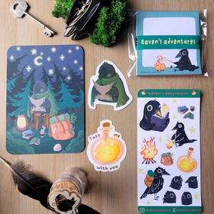 K&ouml;nnte beinhalten: Ein Set aus Schreibwaren mit Raben-Illustrationen, bestehend aus einem Notizblock mit der Aufschrift Raven's adventures, einer Postkarte, zwei Aufklebern, einem Stickerbogen, einer schwarzen Rabenfigur, einem Metallschl&uuml;ssel und einer schwarzen Feder.