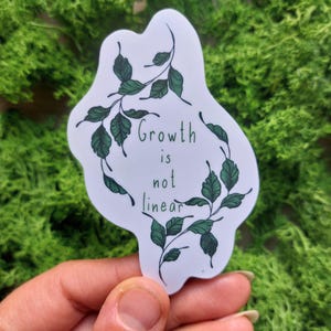 May include: A white sticker featuring the words "Growth is not linear" in green, encircled by a green leafy pattern. The sticker is set against a backdrop of green moss.