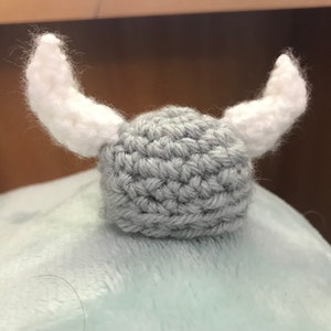 Puede incluir: Un gorro gris de ganchillo con dos cuernos blancos de ganchillo. El gorro está sobre una superficie azul claro y peluda.