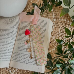Peut inclure: Un marque-page rectangulaire transparent orné d'un motif floral rose, surmonté d'un ruban rose clair. Une chaîne dorée y est fixée, agrémentée d'une breloque fraise rouge, d'un petit cœur doré, d'un cœur rose et d'une breloque coquillage doré.