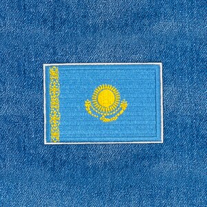 May include: A rectangular patch displaying the flag of Kazakhstan. The flag has a light blue field with a yellow sun and a decorative column. It is framed by a white border, set against a textured blue backdrop.