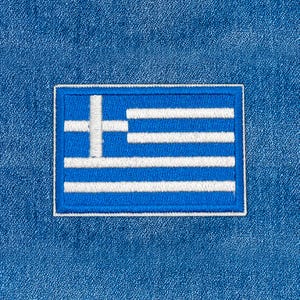 May include: A rectangular patch featuring the flag of Greece, with a white cross in the upper left corner and alternating blue and white horizontal stripes. The patch is bordered by a white outline and set against a blue denim background.