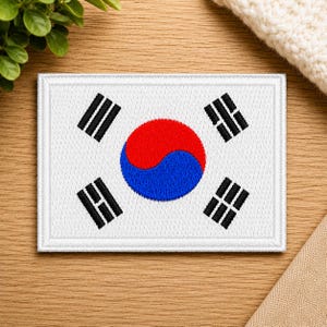 May include: A rectangular embroidered patch featuring the South Korean flag design. The center displays a red and blue taegeuk symbol, surrounded by four black trigrams on a white background, all enclosed within a stitched white border.