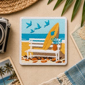 May include: Embroidered patch featuring a beach scene with a yellow surfboard, white bench, and blue birds flying over the ocean. A small vase of flowers sits on the bench. The patch has a white border and is square-shaped.