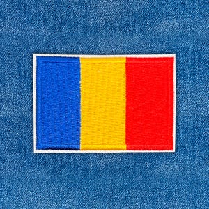 May include: Embroidered patch featuring the flag of Romania. The rectangular patch has vertical stripes in blue, yellow, and red, outlined with a white border. The patch is set against a blue denim background.