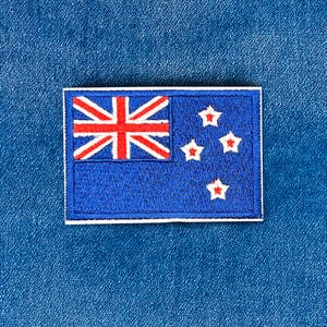 May include: An embroidered patch of the New Zealand flag. The flag is blue with the Union Jack in the top left corner and four red-bordered white stars on the right. The patch is rectangular with a white border.