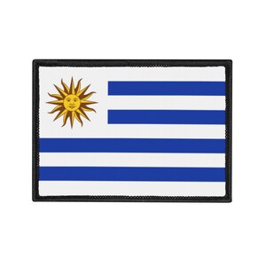 Pode incluir: Um patch retangular com a bandeira do Uruguai. A bandeira tem um fundo branco com um sol dourado no canto superior esquerdo. Existem quatro listras azuis e cinco brancas. O patch tem uma borda preta.