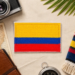 Può includere: Toppa ricamata raffigurante la bandiera della Colombia. La toppa rettangolare mostra strisce orizzontali gialle, blu e rosse, con un bordo bianco. La toppa è su una superficie beige con oggetti a tema viaggio.
