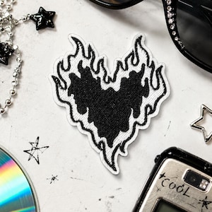 Puede incluir: Un parche bordado con forma de corazón rodeado de llamas estilizadas. El corazón es de color negro sólido, mientras que el patrón de llamas circundante está delineado con hilo blanco. El parche presenta un diseño de alto contraste.