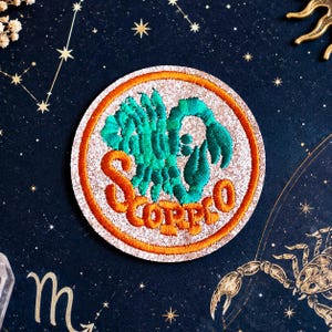 May include: A circular embroidered patch featuring a shimmering silver background with a bright orange border. The centre displays a teal scorpion graphic positioned above the orange embroidered text that reads SCORPIO in a bold, rounded font.