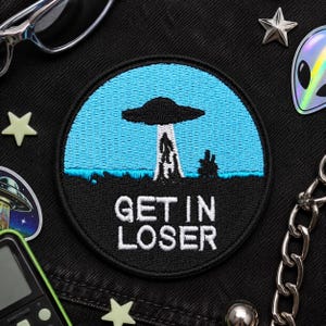 Peut inclure: Écusson rond brodé avec une bordure noire, représentant un ciel bleu et un OVNI noir. L'écusson porte l'inscription "GET IN LOSER" en blanc, avec un paysage en dessous. L'écusson est entouré d'autres accessoires.