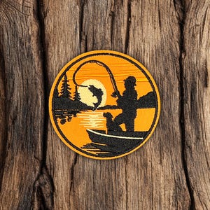 May include: Round embroidered patch featuring a fishing scene. The design includes a fisherman in a boat, a fish jumping, and a sunset over a lake. The patch has an orange and black color scheme and is set against a wooden background.