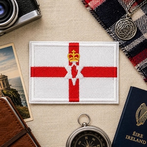 May include: A rectangular embroidered patch featuring the flag of Northern Ireland, which consists of a white field with a red cross, a central six-pointed star, a red hand emblem, and a yellow crown positioned above the red hand symbol.