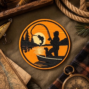 May include: Circular embroidered patch depicting a fishing scene with a fisherman in a boat, a fish leaping, and a sunset. The patch has an orange and black colour scheme. Fishing tackle and a compass surround the patch.