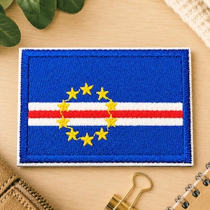 May include: A rectangular embroidered patch featuring the flag of Cape Verde. The design consists of a blue field with a horizontal red stripe bordered by two white stripes, and a circle of ten yellow five-pointed stars centered on the left side.