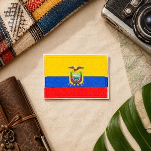 May include: Embroidered patch featuring the flag of Ecuador, with a yellow top, blue center, and red bottom. The flag includes the Ecuadorian coat of arms. The patch is rectangular and has a white border. Other items in the image include a camera, map, and journal.
