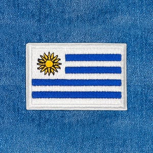 Pode incluir: Patch bordado com a bandeira do Uruguai. O patch retangular tem uma borda e fundo brancos. O design da bandeira inclui um sol amarelo no canto superior esquerdo e listras azuis e brancas.