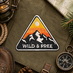 Op de afbeelding: Een driehoekige patch met een berglandschap en de woorden "WILD & FREE". De patch heeft een zwart berg silhouet tegen een verloop van oranje en geel, met een witte rand. De patch is omgeven door outdoor avontuurlijke uitrusting.