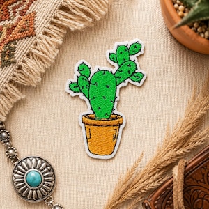 Puede incluir: Parche bordado con un cactus verde en una maceta amarilla. El cactus tiene costuras detalladas y está sobre un fondo neutro. Adecuado para ropa o accesorios.