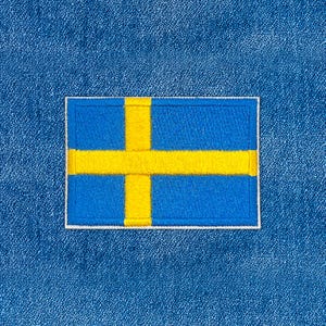 Puede incluir: Un parche rectangular con la bandera de Suecia. La bandera tiene un fondo azul con una cruz escandinava amarilla. El parche está bordeado con una costura blanca y está sobre un fondo de mezclilla azul.