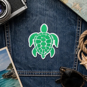 Peut inclure: Un écusson vert en forme de tortue avec une bordure blanche, posé sur une surface en jean bleu. L'écusson présente un motif de tortue détaillé. D'autres éléments dans l'image incluent un appareil photo vintage, des lunettes de soleil, une carte, une carte postale, une corde et une étoile de mer.