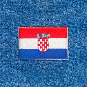 May include: Embroidered patch featuring the Croatian flag. The flag design includes horizontal stripes of red, white, and blue, with the Croatian coat of arms in the center. The patch is rectangular with a white border, set against a blue denim background.