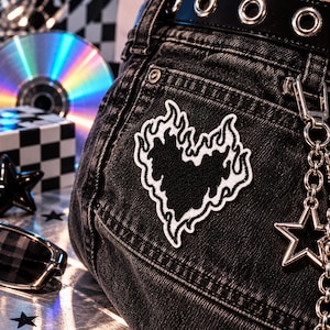 May include: A black denim garment featuring a white embroidered patch of a heart surrounded by flames. The item includes a black belt with silver grommets and a silver metal chain with a star-shaped charm attached to the side of the fabric.