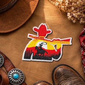 Könnte beinhalten: Gestickter Aufnäher mit einem Cowboy auf einem Pferd, der einen Stier vor einem Sonnenuntergang einfängt. Der Aufnäher ist rot, gelb und schwarz, mit einem weißen Rand. Das Bild ist von einem braunen Cowboyhut, Stiefeln und einem Gürtel umgeben.