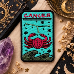 Könnte beinhalten: Ein rechteckiger bestickter Aufnäher mit schwarzem Rand, der oben das Wort CANCER in pinkfarbenem Text zeigt. Das Design umfasst ein grünes Krebs-Sternbild, einen großen pinkfarbenen Krebs und eine dunkelgrüne Welle auf helltürkisem Grund.