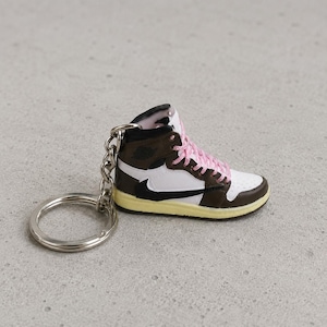 Può includere: Un portachiavi a forma di sneaker alta in miniatura con base bianca, inserti marrone scuro, un logo swoosh rovesciato nero e lacci rosa acceso. La scarpa presenta un'intersuola color crema e un anello portachiavi in metallo argentato sul tallone.
