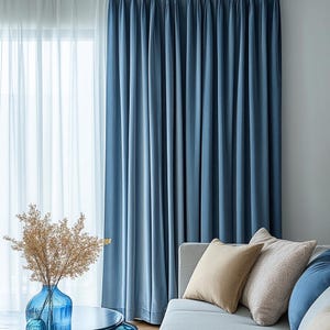 May include: A pair of blue curtains with vertical folds hangs next to a sheer white curtain. A blue glass vase with dried flowers sits on a round table. Beige and blue pillows rest on a gray sofa.