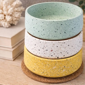 May include: A stack of three speckled ceramic bowls in pastel colors: mint green, white, and yellow. Each bowl rests on a cork coaster. The bowls are stacked on a wooden surface, with a decorative coral and books in the background.