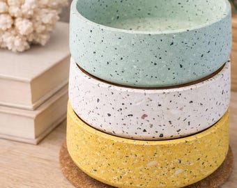 May include: A stack of three speckled ceramic bowls in pastel colors: mint green, white, and yellow. Each bowl rests on a cork coaster. The bowls are stacked on a wooden surface, with a decorative coral and books in the background.
