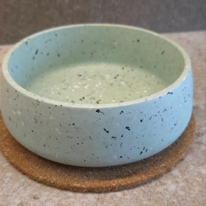 Op de afbeelding: Een ronde, lichtblauwe kom met een gespikkeld ontwerp, rustend op een ronde kurken onderzetter. De kom heeft een dikke, afgeronde rand en een glad interieur. Het gespikkelde ontwerp bevat zwarte en witte vlekken.
