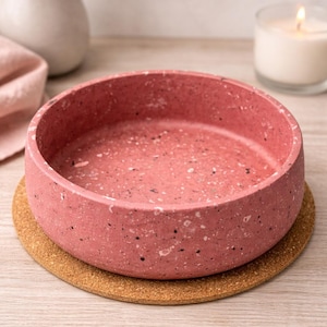 Pode incluir: Uma tigela redonda rosa com um design salpicado assenta sobre um descanso de copo redondo de cortiça. A tigela é feita de um material sólido e tem um interior liso. Uma vela acesa e um vaso com flores secas estão no fundo.