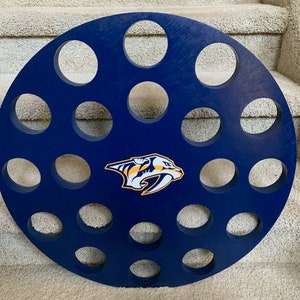 May include: A round, blue wooden beer can holder with 20 holes for cans. The holder features a Nashville Predators logo in the center.