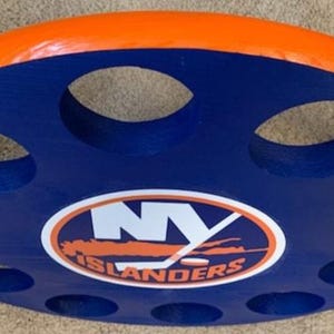 May include: A round, wooden drink holder with a blue base and orange trim. The holder features multiple circular cutouts and the New York Islanders logo in the center. A decorative item for hockey fans.