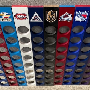 May include: A set of 10 wooden hockey puck holders, each featuring a different NHL team logo. The holders are painted in various colors, including blue, red, white, black, and purple. Each holder has 10 circular cutouts for holding pucks.
