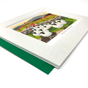 May include: A white card with a green envelope. The card features a colorful illustration of a group of sheep standing in a line, with a red gate in the background. The text "Sorry I'm so Late" is printed on the card.