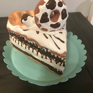 May include: A slice of cake decorated with chocolate frosting, white icing, and a chocolate-covered donut with white icing. The cake is on a light blue cake stand.