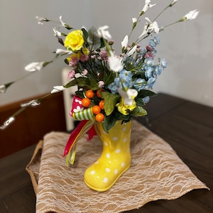 May include: A yellow rain boot with white polka dots filled with a floral arrangement. The arrangement includes yellow roses, blue flowers, white flowers, and green leaves. The boot is sitting on a brown burlap table runner.