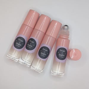 Può includere: Quattro flaconi di profumo roll-on con tappi ed etichette rosa. Le etichette riportano "Dream of Opaline perfume" in bianco su sfondo nero. Un flacone ha il tappo rimosso, rivelando una sfera a rullo argentata.