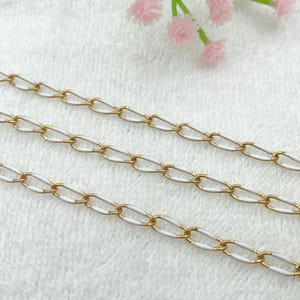 Könnte beinhalten: Nahaufnahme von drei goldfarbenen Kettengliedern mit weißen Emaille-Gliedern. Die Ketten sind diagonal über einer weißen, strukturierten Oberfläche angeordnet. Rosa Blüten sind im Hintergrund zu sehen.