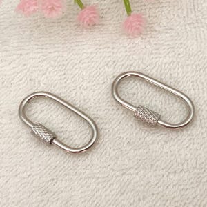 Könnte beinhalten: Zwei silberfarbene ovale Karabinerhaken mit strukturierten Schraubverschlüssen. Sie werden auf einer weißen, strukturierten Oberfläche mit rosa Blütenakzenten gezeigt. Diese Clips eignen sich für verschiedene Bastel- und DIY-Projekte.