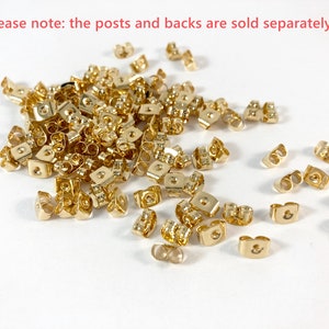 Op de afbeelding: Een grote stapel gouden oorbelruggen. De ruggen zijn van metaal en hebben een kleine lus aan de bovenkant. De tekst "please note: the posts and backs are sold separately" is zichtbaar bovenaan de afbeelding.