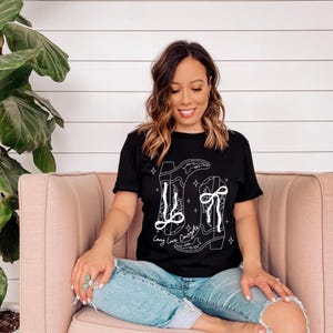 May include: Black t-shirt with a white graphic of cowboy boots and the text "Long Live Cowgirls". The shirt is made of a soft material and is suitable for casual wear. The model is wearing blue jeans.
