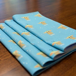 May include: A stack of four blue fabric napkins with a repeating pattern of a cartoon character with the text "Pretty" printed on each napkin.