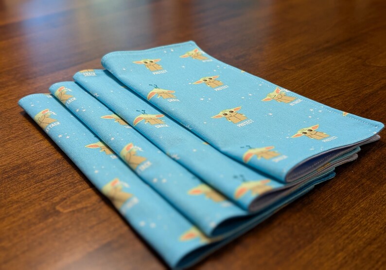 May include: A stack of four blue fabric napkins with a repeating pattern of a cartoon character with the text "Pretty" printed on each napkin.