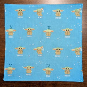 May include: A blue fabric square with a repeating pattern of a green character with different expressions. The character is surrounded by white stars. The text "PROTECT", "ATTACK", and "SNACK" appears on the fabric.